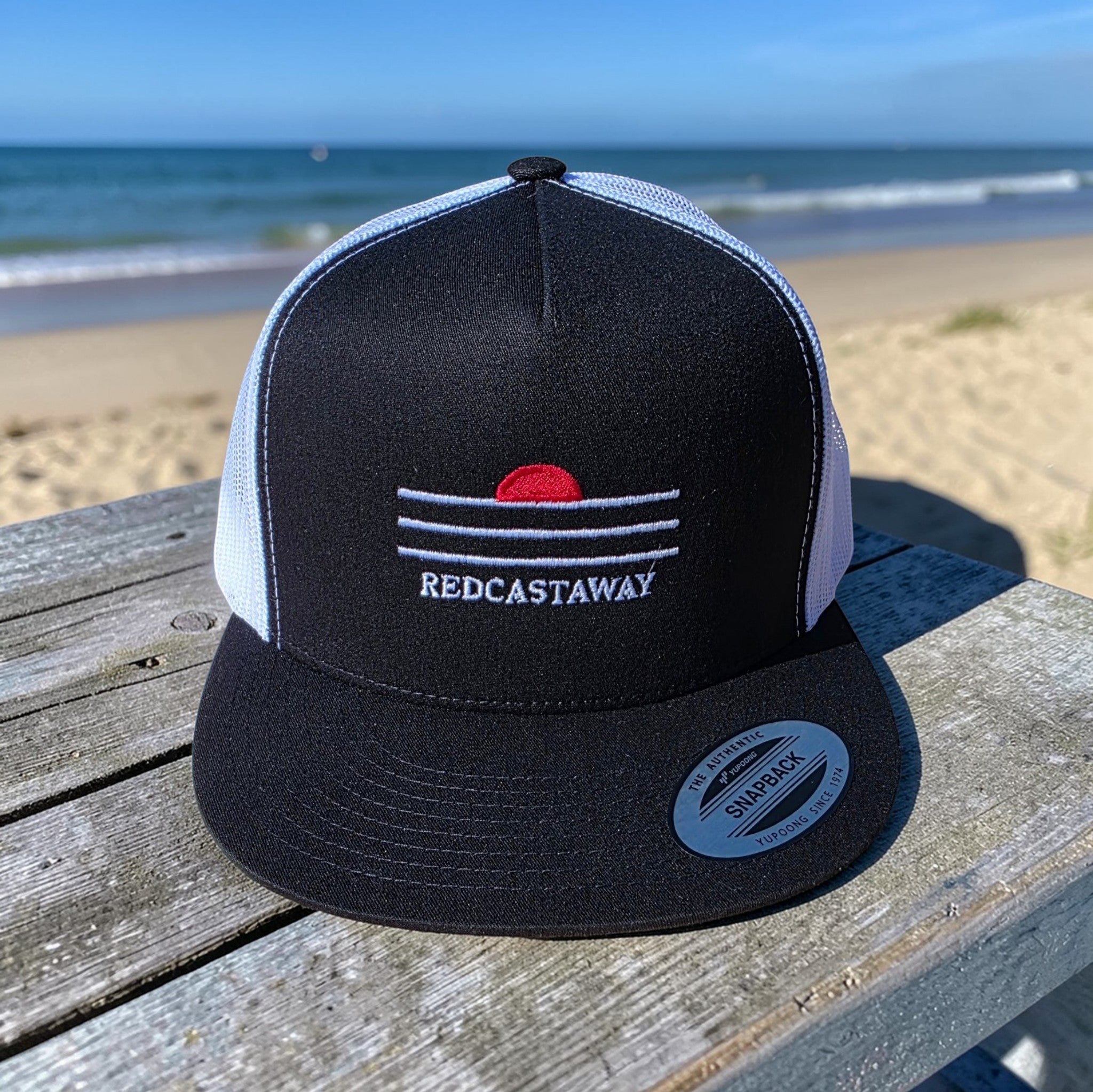 Redcastaway black trucker cap with white mesh back and embroidered sunset logo. Features a flat bill and snapback closure.