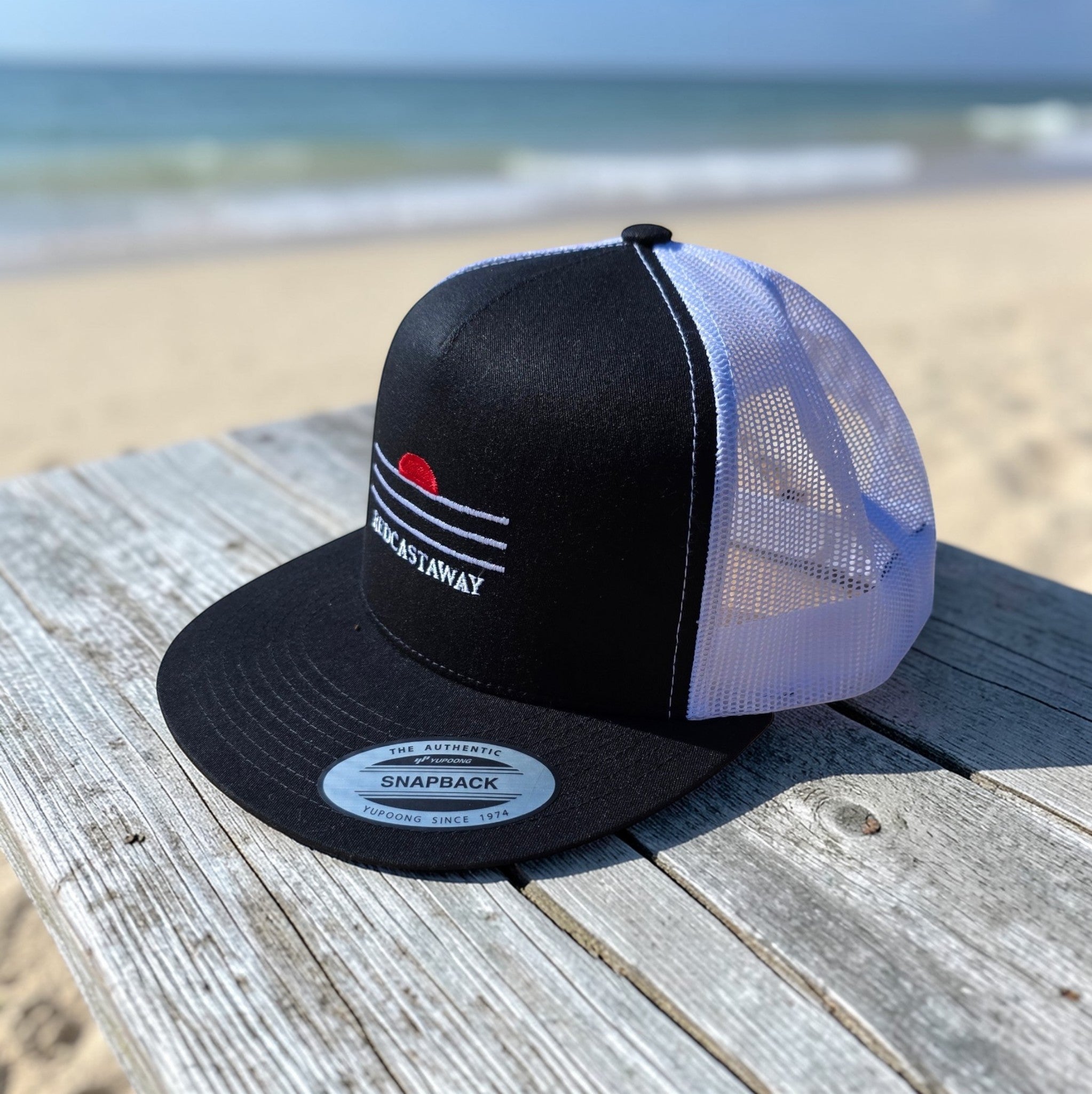 Redcastaway black trucker cap with white mesh back and embroidered coastal logo. Flat bill, snapback closure.