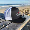 Redcastaway black trucker cap with white mesh back and embroidered logo, flat bill, beach background